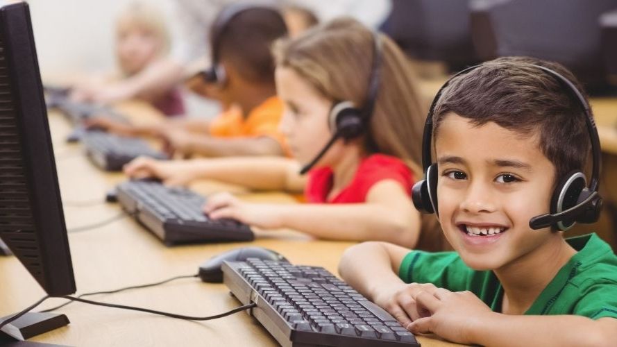 Row of happy children, with headphones on, using desktop computers in a classroom.